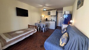 Ett kompakt bostadsutrymme med ett litet kök, en matplats, en soffa med mönstrade kuddar och en TV som är monterad på väggen.