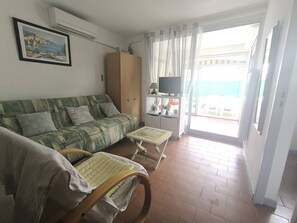 A living room with a sofa, two wooden chairs, a television, and a balcony with a view. - T2 apartment with AC - 4 guests (Cap d'Agde)