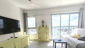 Un soggiorno con TV, divano, tavolino da caffè, un armadietto giallo e una grande finestra vista porto turistico.