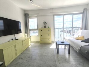 A living room with a TV, a sofa, a coffee table, a yellow cabinet, and a large window with a view of a marina. - Apartment with port view - 6 guests (Cap d'Agde)