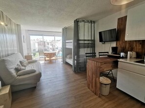 Ein modernes Wohnzimmer mit einer Couch, einem Holzesstisch und einer Küchenzeile inklusive Fernseher und Kaffeemaschine.