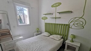 A bedroom with a bed, a window, a bedside table, a nightstand, and a wall with a green design. - Family apartment near the pines - 6 guests (Cap d'Agde)