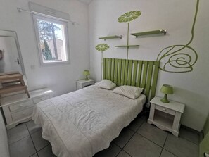 A bedroom with a bed, a window, a bedside table, a nightstand, and a wall with a green design. - Family apartment near the pines - 6 guests (Cap d'Agde)