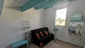 A room with a black sofa, a teal bench, a metal shelf unit, a standing fan, a mirror, and a window with a view. - Family apartment near the pines - 6 guests (Cap d'Agde)