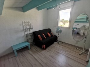 A room with a black sofa, a teal bench, a metal shelf unit, a standing fan, a mirror, and a window with a view. - Family apartment near the pines - 6 guests (Cap d'Agde)