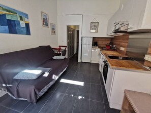 Ein kompakter Wohnbereich mit einer Couch, einer kleinen Küche inklusive Kühlschrank, Waschmaschine und einer Holz Arbeitsplatte.