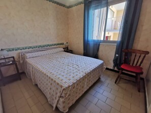 Ein Schlafzimmer mit Bett, einem Sessel, einem Nachttisch, einem Fenster mit Vorhängen und Blick auf einen Balkon.