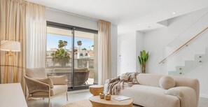 Ein modernes Wohnzimmer mit einer Couch, einem Sessel und einem Couchtisch, einer Treppe und Blick auf Gebäude und Palmen durch große Fenster.