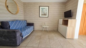 A modern living room with a sofa, a small round mirror, a framed picture on the wall, a television, and a white side table. - Studio cabin sea view, parking - 4 guests (Cap d'Agde)