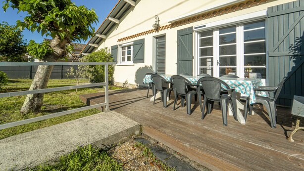 Un patio con mesa y sillas, una terraza de madera y una casa con tejado de tejas y persianas.
