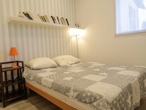 Ein Schlafzimmer mit Bett, Kissen, einem Nachttisch, einer Lampe, einem Bücherregal und einem Fenster.