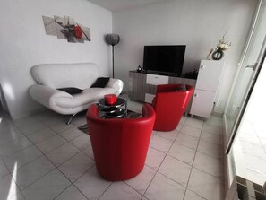Ein modernes Wohnzimmer mit einem weißen Sofa, einem roten Sessel, einem Fernseher und einer Küchenzeile.