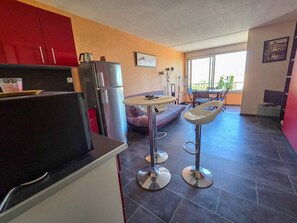 Eine moderne Küche mit Kühlschrank, ein Essbereich mit rundem Tisch und Stühlen sowie ein Blick ins Freie durch große Fenster.