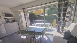 Una habitación iluminada por el sol con una mesa y sillas de comedor, un sofá, un televisor y un gran ventanal corredizo que da a un patio exterior.