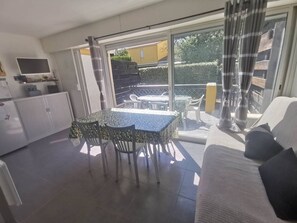 A sunlit room with a dining table and chairs, a sofa, a TV, and a sliding glass door leading to an outdoor patio. - Air-conditioned apartment, garden - 4 guests (Cap d'Agde)