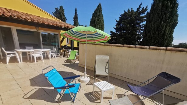 Un patio con mobiliario exterior, incluyendo sillones y una mesa, bajo un paraguas a rayas.
