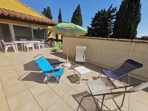 A patio with outdoor furniture, including lounge chairs and a table, under a striped umbrella. - Apartment with AC, pool, near beaches - 8 guests (Cap d'Agde)