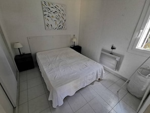 A bedroom with a bed, two bedside tables, a radiator, and a window. - Apartment with AC, pool, near beaches - 8 guests (Cap d'Agde)