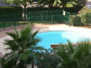 A swimming pool surrounded by a green fence, palm trees, and a hedge. - Apartment with AC, parking - 4 guests (Cap d'Agde)