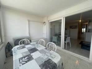 A dining area with a checkered tablecloth, white chairs, and a sliding glass door leading to a kitchen and dining space. - Apartment with AC, parking - 4 guests (Cap d'Agde)