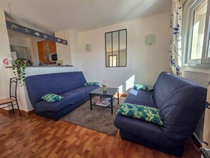 Ein Wohnzimmer mit einer Couch, einem Couchtisch und einem kleinen Beistelltisch. Im Hintergrund ist eine Küchenzeile erkennbar.
