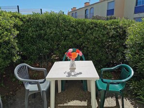 A patio with a table and chairs, a vase with flowers, and a hedge in the background. - Cozy studio near the beach - 3 guests (Cap d'Agde)