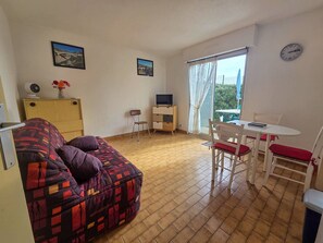 A furnished room with a sofa, a small table with chairs, a TV, and a window with curtains. - Cozy studio near the beach - 3 guests (Cap d'Agde)