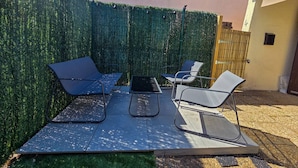 Eine Terrasse im Freien mit Tisch und vier Stühlen, ein grüner Heckenstreifen und ein Gebäude im Hintergrund.