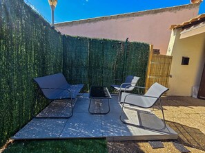Eine Terrasse im Freien mit Tisch und vier Stühlen, ein grüner Heckenstreifen und ein Gebäude im Hintergrund.
