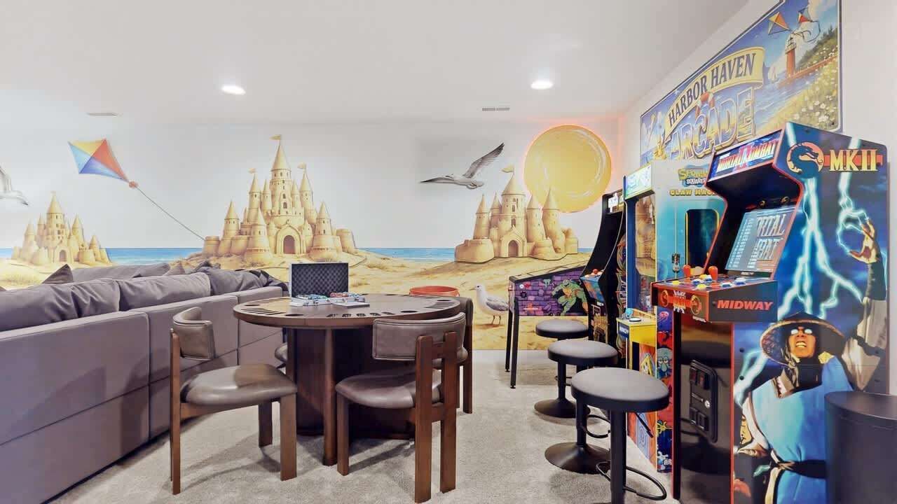 A room with a beach-themed mural, a sofa, a round table with chairs, and arcade games.