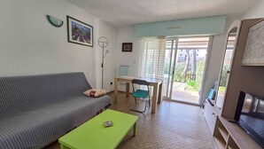 Ein Wohnzimmer mit Sofa, einem grünen Couchtisch, einem Esstisch mit Stühlen, einem Fernseher und einer Glastür, die zu einem Außenbereich führt.