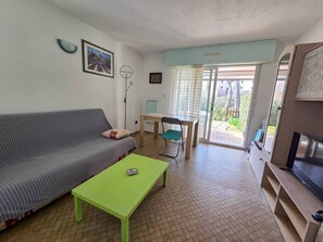 Ein Wohnzimmer mit Sofa, einem grünen Couchtisch, einem Esstisch mit Stühlen, einem Fernseher und einer Glastür, die zu einem Außenbereich führt.