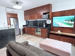 Una cocina moderna con horno empotrado, microondas y fregadero. En la sala de estar hay un sofá gris y un otoman rosa.