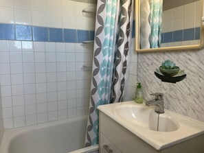 A bathroom with a white sink, a mirror, a shower curtain with a geometric pattern, a small plant on a shelf, and a tiled wall with a blue stripe. - 2-room apartment sea view - 4 guests (Cap d'Agde)