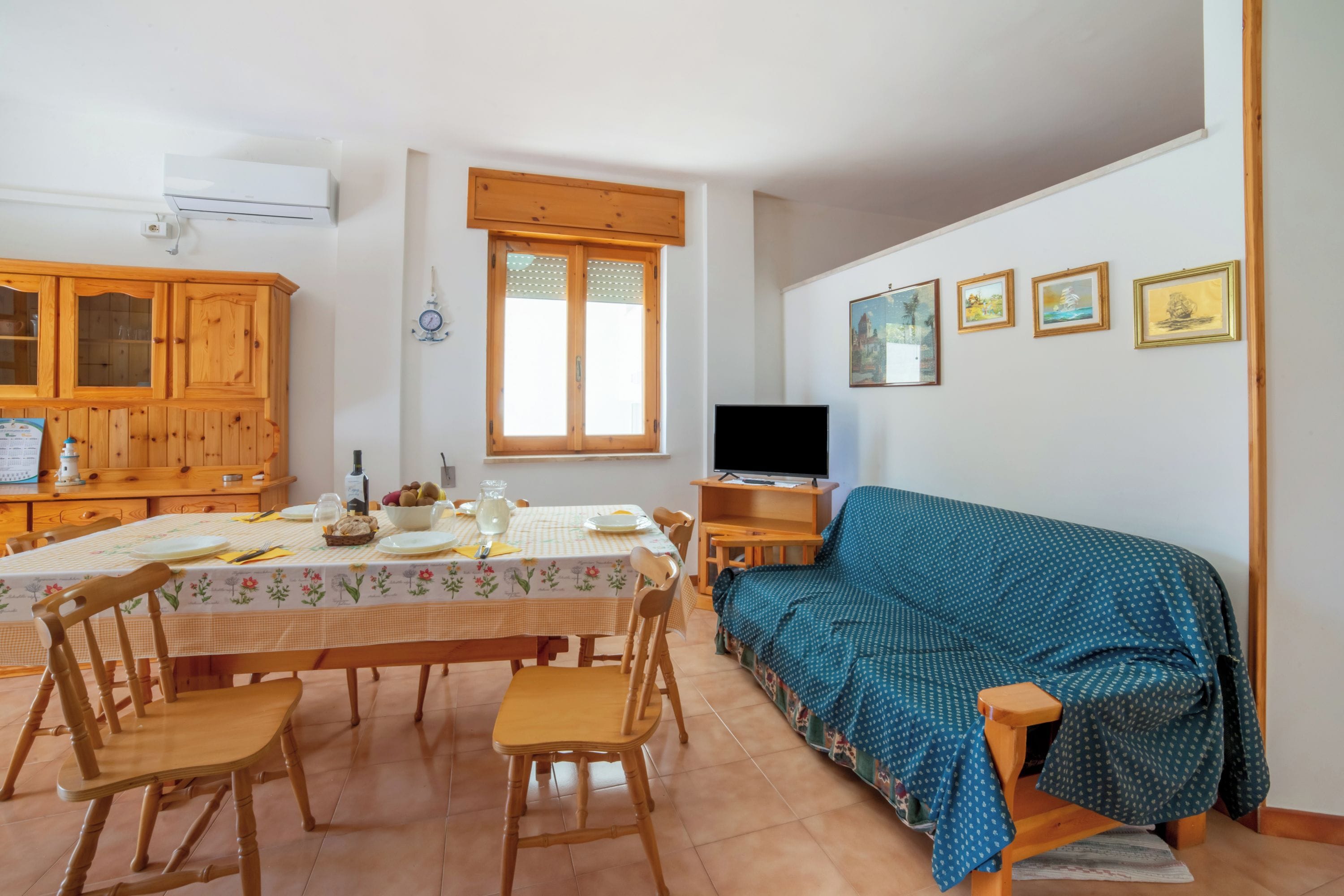 Uma sala de jantar com mesa posta para quatro pessoas, um armário de madeira, uma televisão e um sofá com capa estampada.