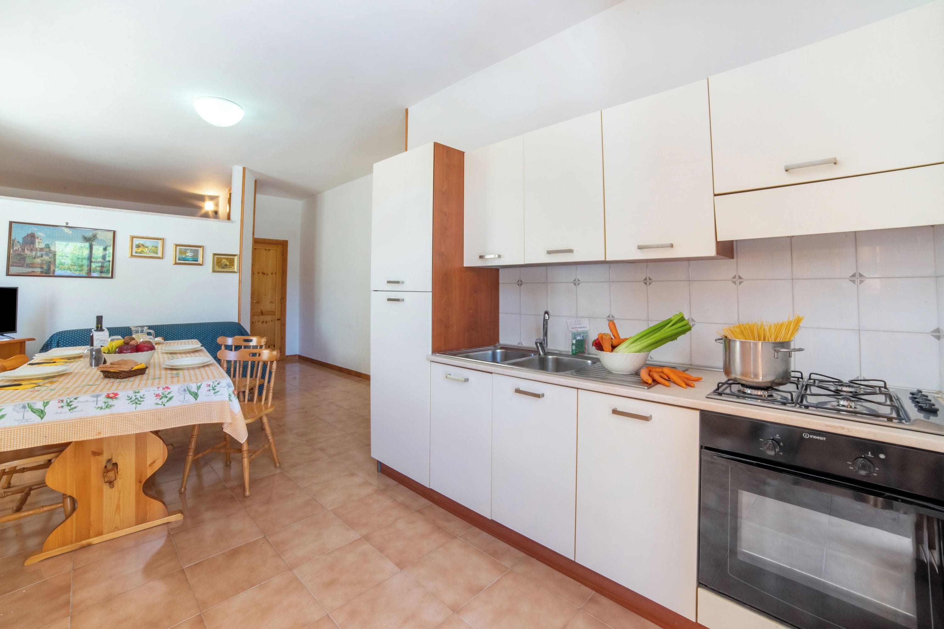 Cozinha com armários brancos, fogão e pia. Há uma mesa de jantar com cadeiras e toalha de mesa.