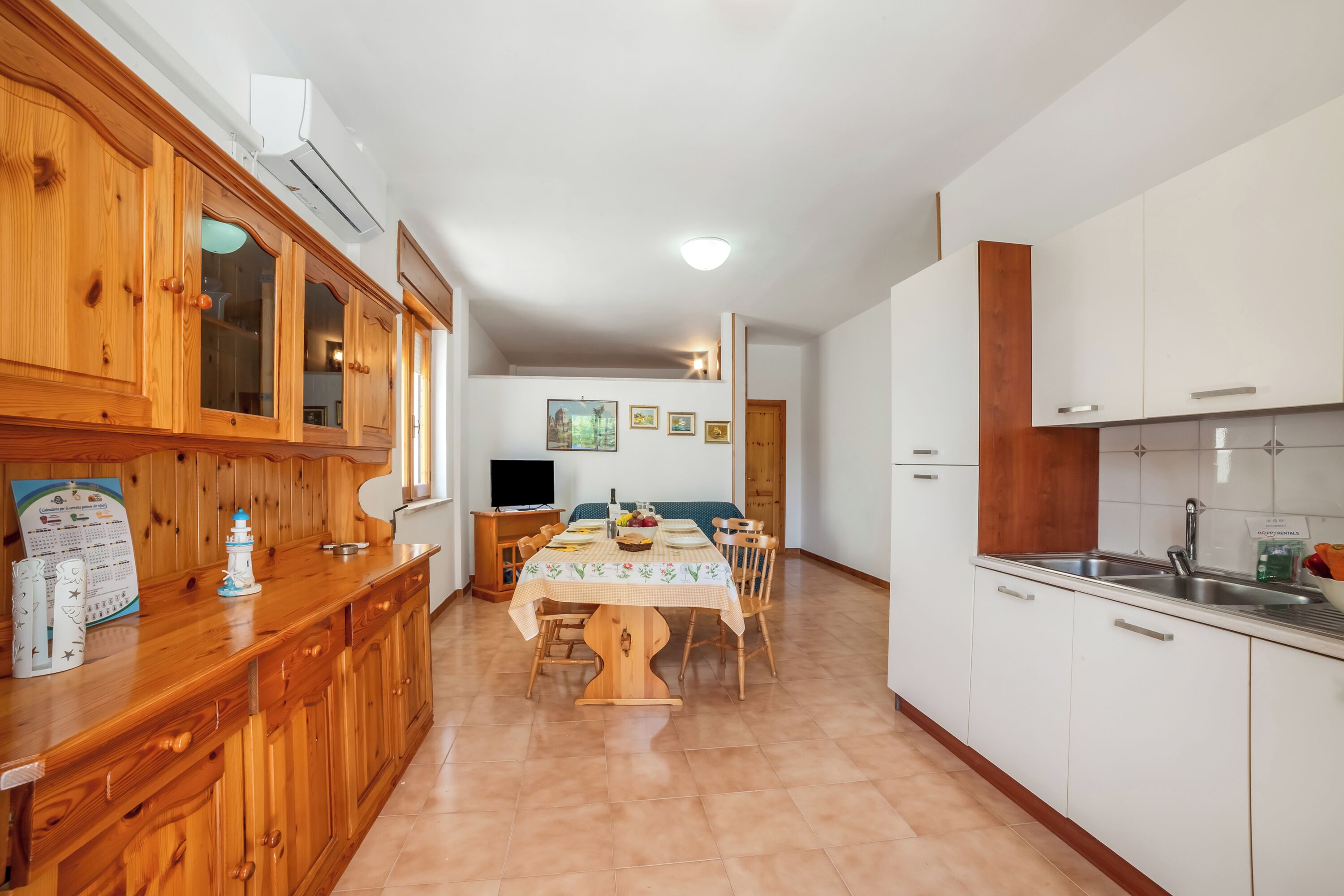 Uma cozinha com armários de madeira, uma mesa de jantar com cadeiras, uma televisão e uma geladeira.