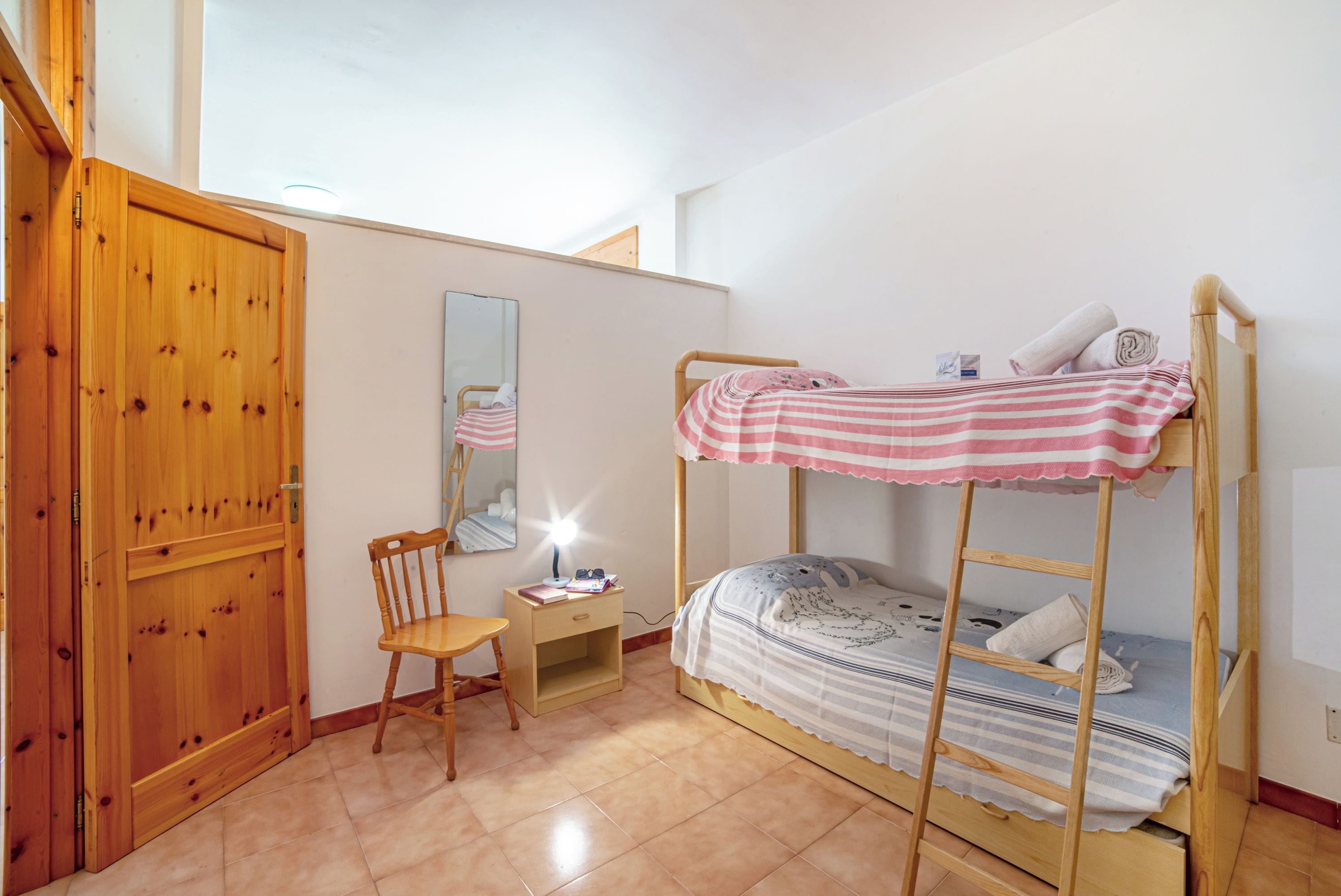 Una cama litera con colcha a rayas, un escritorio de madera con silla, un espejo y un armario de madera.