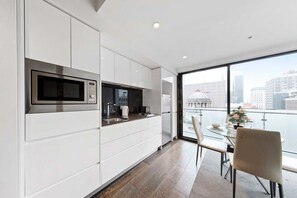 Eine moderne Küche mit integriertem Mikrowellenherd, einer Kaffeemaschine und einem Spülbecken. Ein Essbereich mit Tisch und Stühlen lädt zum Verweilen ein, und ein großes Fenster bietet Blick auf die Stadt.