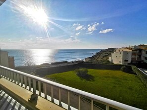 Blick aufs Meer vom Balkon mit Grünfläche, felsigem Ufer und Gebäuden im Hintergrund.