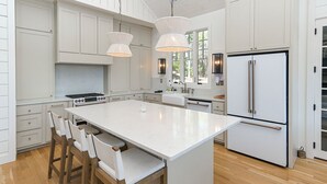 A spacious kitchen with white cabinetry, a large island, wooden chairs, and a stainless steel refrigerator. - Luxurious High End Family Friendly Lake House. Dock, Floats, 10 min to Trident. (Crane Hill)