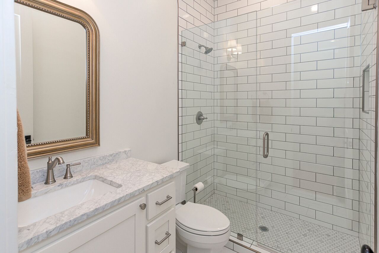A bathroom with a marble countertop, a white toilet, a glass-enclosed shower, and a gold-framed mirror.