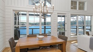 A dining area with a wooden table, chairs, and a chandelier. Large windows offer a view of the water and trees. - Luxurious High End Family Friendly Lake House. Dock, Floats, 10 min to Trident. (Crane Hill)