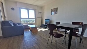 Een moderne woonkamer met een bank, een eettafel met stoelen, een televisie en een groot raam.