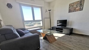 Een moderne woonkamer met een grijze bank, een glazen salontafel, een tv op een meubelstuk, en een groot raam met uitzicht op de omgeving.