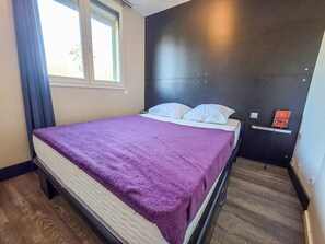 A bedroom with a bed, a window, a dark headboard, and a small shelf with an orange item. - Air-conditioned apartment in Môle - 4 guests (Cap d'Agde)