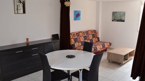 A dining area with a round table, black chairs, a dark-colored cabinet, and a colorful sofa. - Quiet apartment with parking - 6 guests (Cap d'Agde)