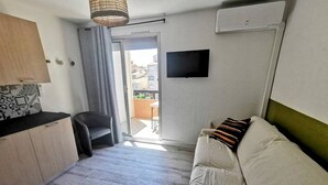 Quarto compacto com sofá, cadeira, mesa pequena, TV e ar condicionado.