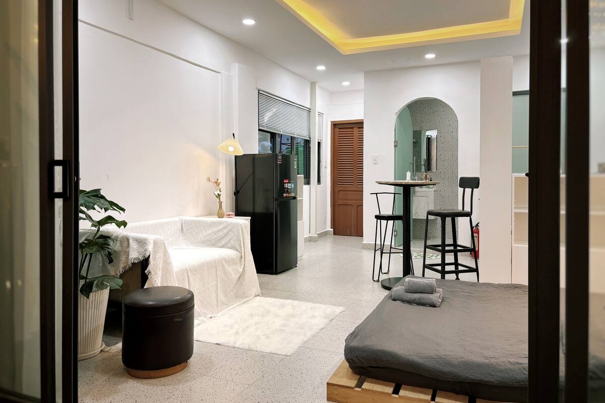 A modern hotel room with a bed, a small table, a chair, a refrigerator, a potted plant, and a bathroom visible through an archway.
