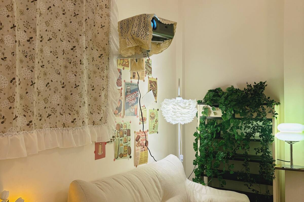 A cozy living room with a white sofa, a ceiling fan, a wall-mounted air conditioner, a decorative lamp, and a wall adorned with various posters.